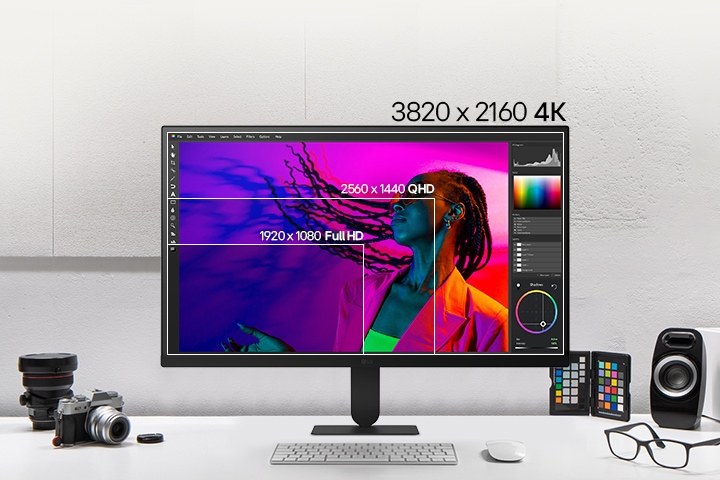Front view of a white LG monitor on a desk, displaying layered screen resolutions for comparison, with cameras, books, color chips, and speakers arranged around it.