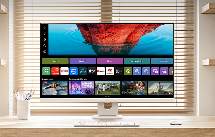 明るいホームオフィスの清潔な木製デスクに設置されたLG SMART Monitor。画面にはwebOSホーム画面が表示され、ストリーミングアプリやコンテンツタイルが配置されている。キーボードとマウスが接続され、背景には本棚とブラインドが映り込む。スマートなエンターテインメントと生産性向上の活用シーンを強調した構図。