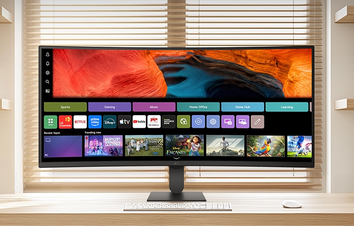 明るいホームオフィスにある清潔な木製デスクに置かれたLGスマートモニター。webOSのホーム画面にはストリーミングアプリケーションとコンテンツタイルが表示され、キーボードとマウスがペアリングされています。背景には本棚とブラインドがあり、スマートなエンターテイメントと生産性のある利用を強調しています。