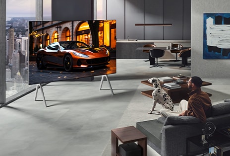 Гостиная с LG OLED TV и звуковой панелью LG Soundbar, установленными на стене. Приставка для нулевого подключения едва заметна под боковым столиком рядом с диваном, что показывает, как легко ее можно разместить в любом месте. Вся планировка гостиной выглядит аккуратно и опрятно. 