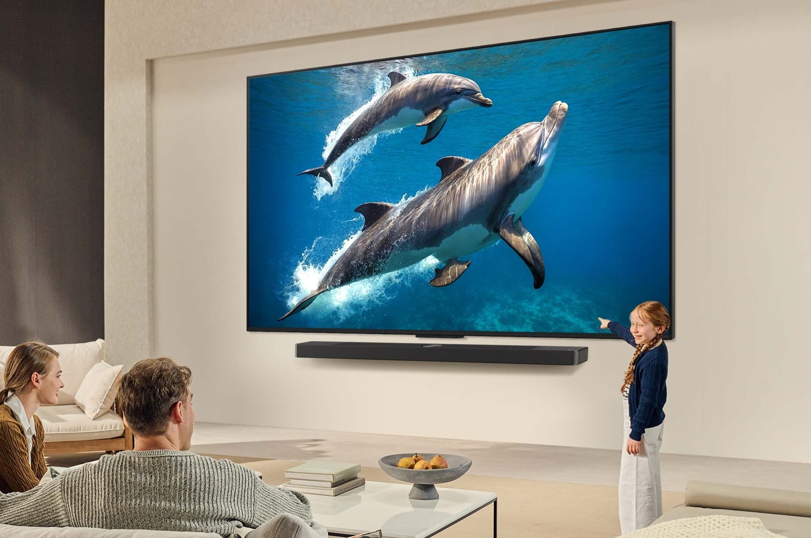 Семья сидит на диване лицом к установленному на стене телевизору LG QNED TV, а сверху звуковая панель LG Soundbar с маленькой девочкой, указывающей на экран, где изображены два дельфина. Заголовок рассказывает о том, как каждое действие становится более захватывающим на огромном экране.