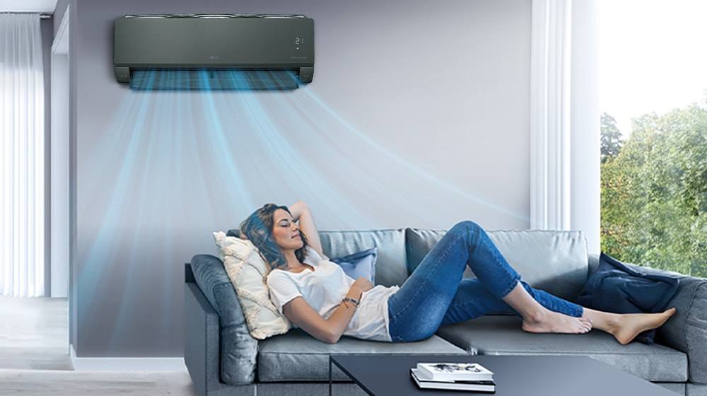 Женщина отдыхает на диване в гостиной, над ней на стене установлен кондиционер LG. Синие потоки воздуха на изображении указывают на то, что он включен и охлаждает комнату.