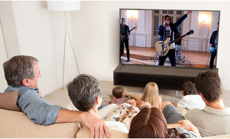 Семья из семи человек находится в гостиной и смотрит фильм. На экране выступает группа музыкантов.