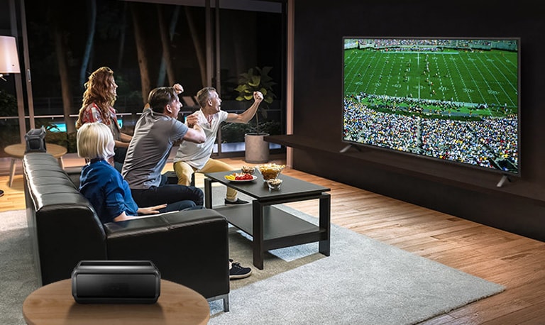 Люди смотрят спортивную игру в гостиной с помощью подключенных через Bluetooth тыловых колонок.