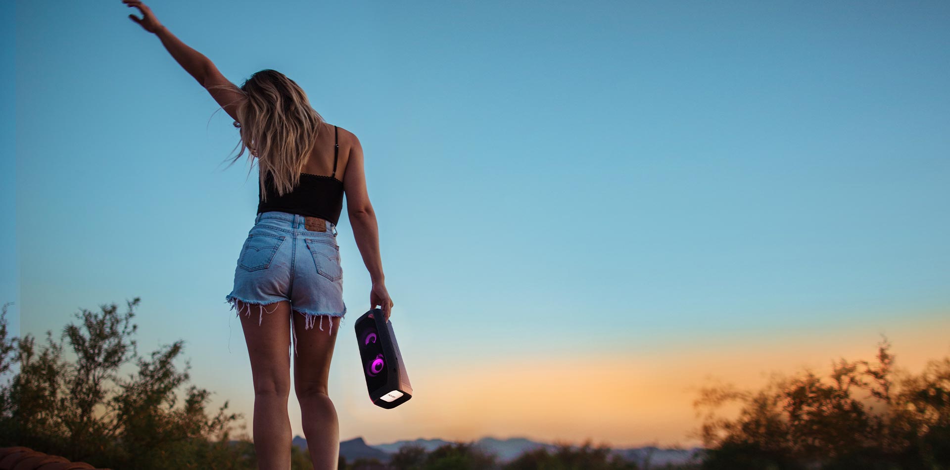 Girl with her back turned is holding a LG Xboom standing in an open field near sunset.