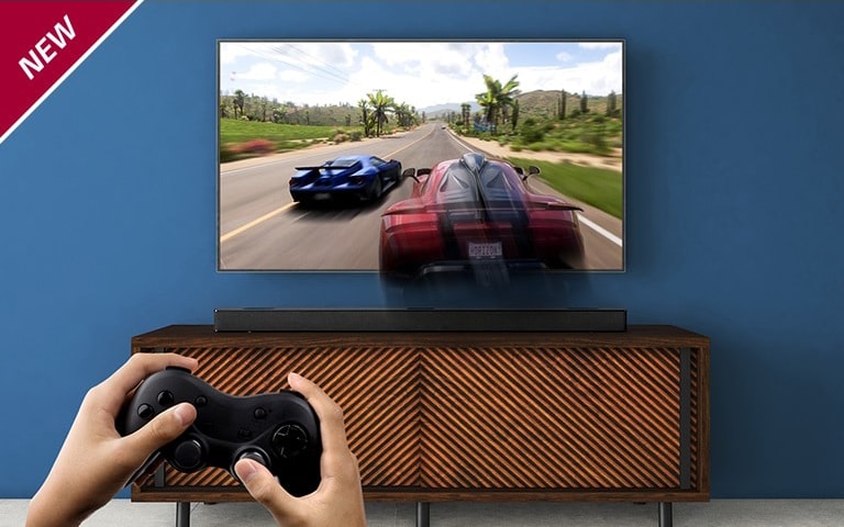 На телевизоре LG, закрепленном на стене, демонстрируется гоночная игра. Саундбар LG расположен на коричневой полке, прямо под телевизором LG. Мужчина держит в руках геймпад. НОВАЯ маркировка показана в верхнем левом углу.