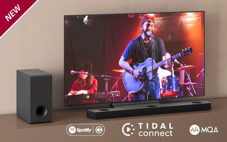 Телевизор LG стоит на серой полке, перед телевизором стоит саундбар LG S95QR. Слева от телевизора стоит сабвуфер. На телевизоре демонстрируется сцена концерта. НОВАЯ маркировка показана в верхнем левом углу.
