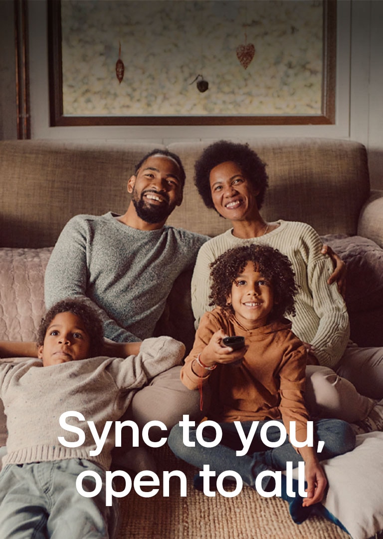 Изображение семьи из четырех человек, сидящей на полу перед уютным коричневым диваном, смотрящей прямо перед собой и смеющейся во время просмотра телевизора. Над изображением белым шрифтом напечатаны слова «Syncs to you, open for all». 