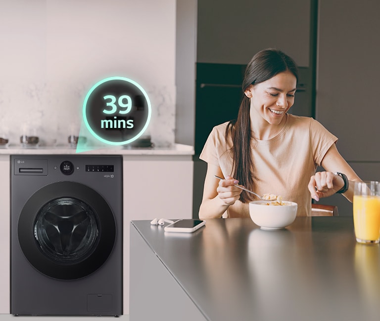 La femme est assise à la table et regarde sa montre, et il y a une machine à laver derrière elle.