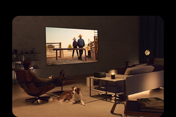 Un hombre viendo una película de vaqueros con su perro en una acogedora sala de estar por la noche. No hay cables alrededor del televisor LG OLED montado en la pared, y la Wireless Connect Box se ubica ordenadamente sobre una mesa auxiliar. Y luego, la imagen se desliza a otra imagen que muestra a un hombre viendo una película de vaqueros con el Televisor LG OLED sobre un soporte de suelo en un espacio acogedor y oscuro. No hay cables alrededor del televisor LG OLED y su soporte de piso, y la Wireless Connect Box se ubica perfectamente sobre una mesa auxiliar