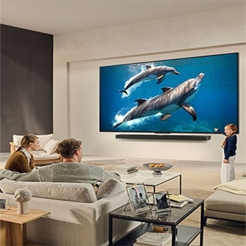 Una familia sentada en el sillón frente a la LG QNED TV montada en la pared encima de una LG Soundbar; una pequeña niña señala la pantalla que muestra dos delfines. El título habla de cómo las escenas son más emocionantes en una pantalla gigante.