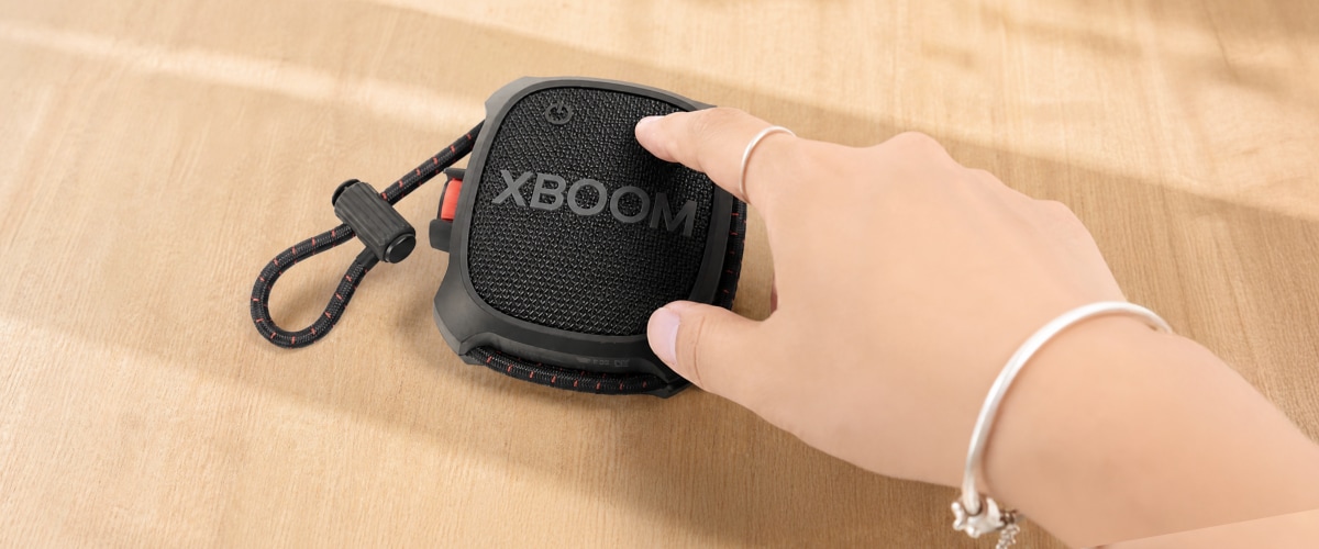 Una mano alcanzando la bocina LG xboom sobre una mesa de madera, con unos audífonos a un lado