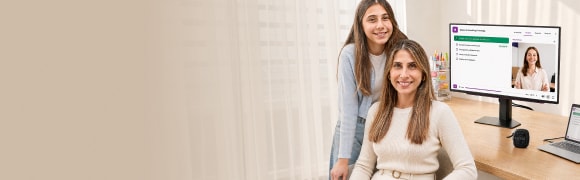 Un banner panorámico con espacio a la izquierda que muestra a una madre y su hija adolescente sonriendo juntas en un estudio en casa: la madre está sentada en primer plano mientras su hija se apoya a su lado. Al fondo, sobre una mesa de madera, se observa un monitor con un curso en línea y una llamada de video, junto con un pequeño altavoz.