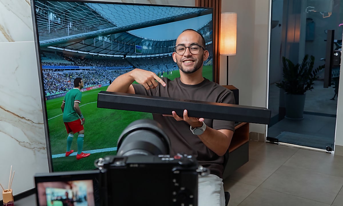 Rapael sonriendo y señalando una barra de sonido que sostiene, con una TV mostrando un partido de fútbol en el fondo