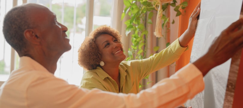 Sandra cuelga una planta en la pared naranja mientras Manuel la ayuda.