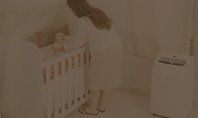 Una madre se inclina sobre la cuna para atender a su bebé en una habitación luminosa y acogedora, con un aire acondicionado portátil LG de color blanco junto a la ventana.