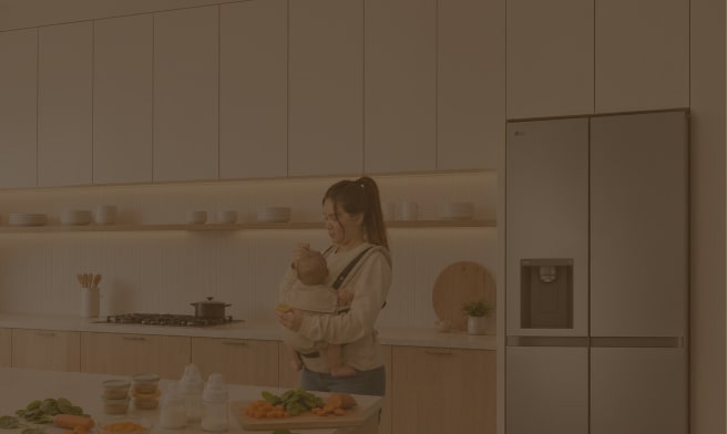 Una madre que lleva a su bebé en un portabebés prepara alimentos en una cocina moderna y luminosa, junto a un refrigerador LG.