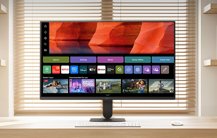 LG Smart Monitor on a clean wooden desk in a bright home office, displaying the webOS home screen with streaming apps and content tiles, paired with keyboard and mouse, bookshelf and window blinds in the background, highlighting smart entertainment and productivity use.