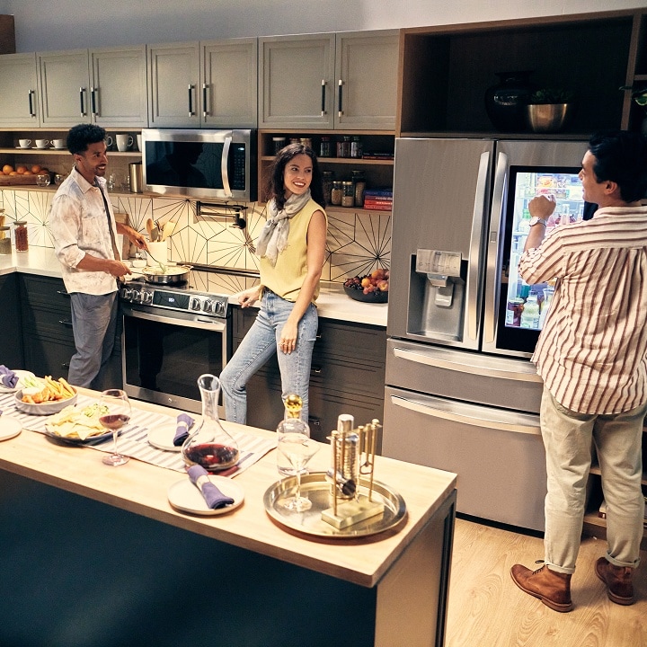 Amigos disfrutando de la comida y la conversación en una cocina moderna con un refrigerador LG de dos puertas en la parte superior y un cajón inferior, mientras uno de ellos revisa los artículos del interior y los demás preparan y comparten la comida juntos.
