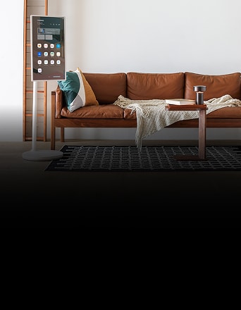 LG StanbyME, una pantalla inteligente vertical, se encuentra junto a un sofá de cuero en una sala de estar moderna, mostrando íconos y widgets de aplicaciones en la pantalla.