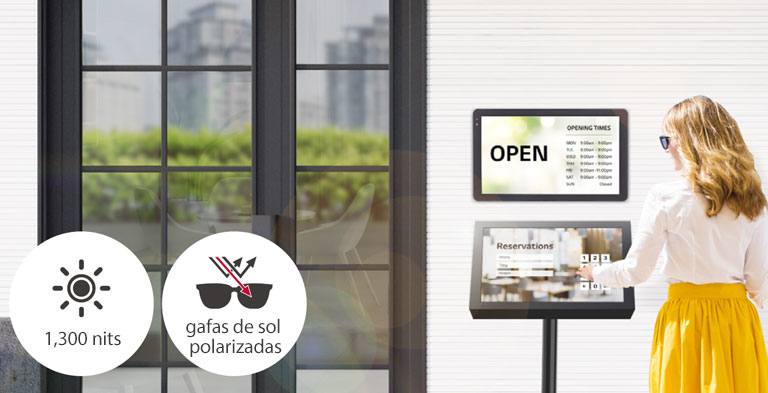 Se instala un 22XF1TJ en la pared de la entrada del edificio con otro soporte 22XF1TJ debajo para fines de reserva. La señora de las gafas de sol está utilizando la pantalla táctil para hacer una reserva. La pantalla permanece visible incluso bajo la luz solar directa.