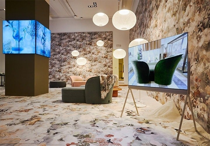Una habitación elegante y moderna con una decoración de temática floral con paredes y alfombras estampadas. Varios televisores LG OLED muestran imágenes vívidas, incluido un primer plano de sillas verdes. La habitación incluye muebles contemporáneos, como una silla rosa y un sofá verde, bajo luces colgantes suaves y esféricas.