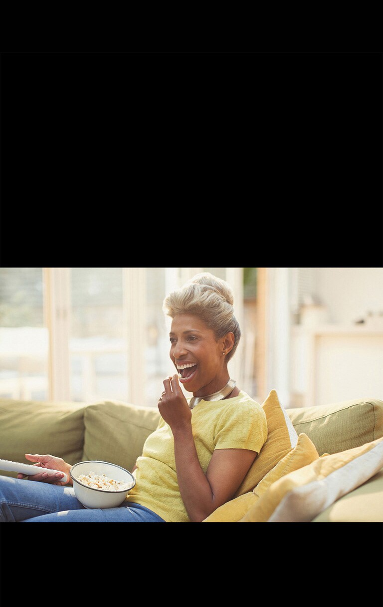 Una mujer está viendo la televisión, y sostiene un control remoto. También está comiendo palomitas de maíz.