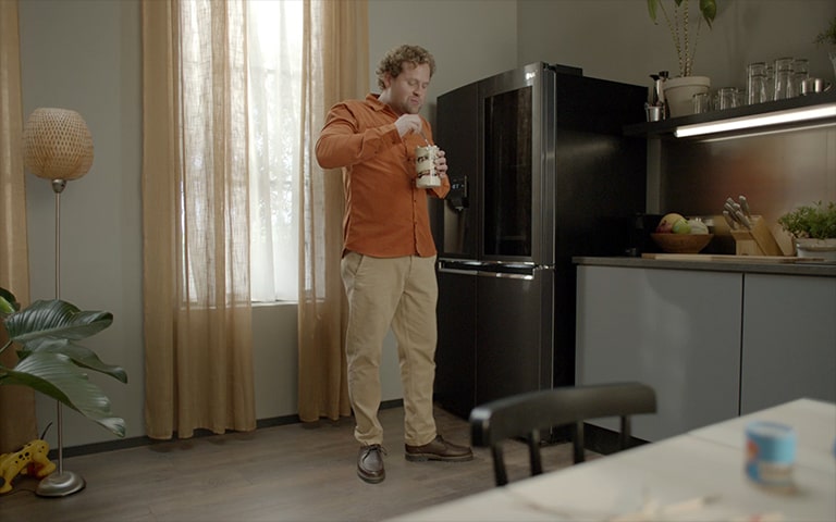 Un hombre de pie junto al refrigerador mientras come un pastel. Esta imagen cambia para mostrar otra imagen. Un hombre toca la puerta del refrigerador y se puede ver lo que hay dentro.