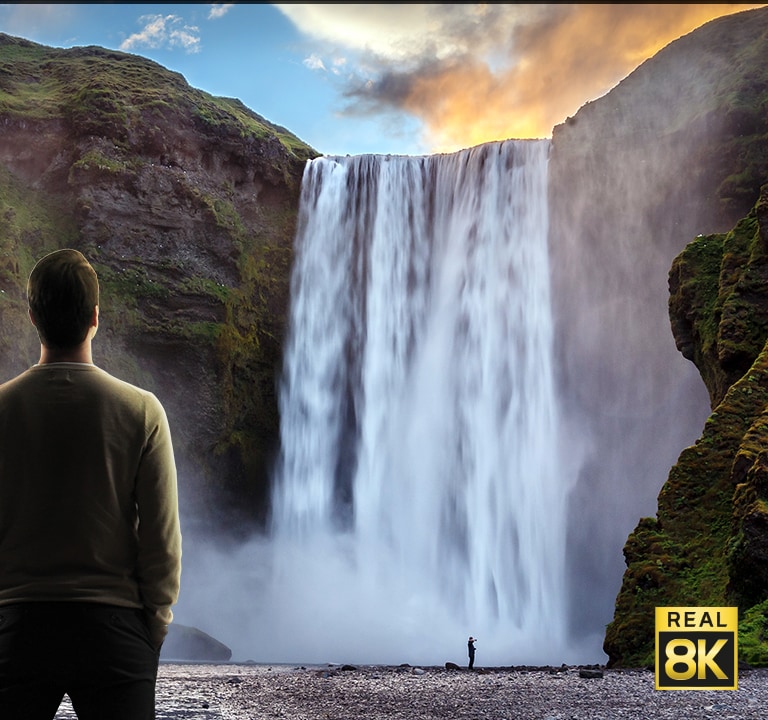 Un hombre se paró frente a una imponente vista de una gran cascada que se precipitaba por los acantilados. La escena se aleja para mostrar la cascada como una imagen en un televisor montado en la pared.