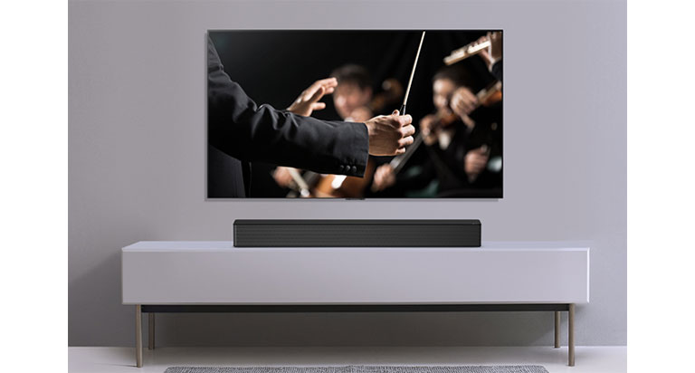 Se muestra un televisor en una pared gris y LG Soundbar debajo de él en un estante gris. La televisión muestra a un conductor dirigiendo una orquesta.