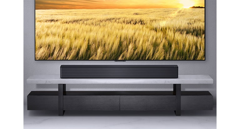 Se muestra un televisor en una pared gris y LG Soundbar debajo de él en un estante gris. Disco Blue-Ray debajo del estante.