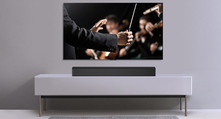 Se muestra un televisor en una pared gris y LG Soundbar debajo de él en un estante gris. La televisión muestra a un conductor dirigiendo una orquesta.