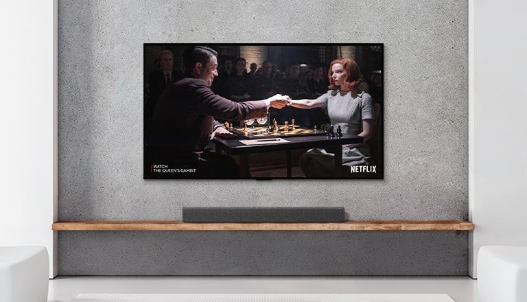 Una barra de sonido y TV están en una sala blanca. Una mujer y un hombre juegan al ajedrez en la pantalla del televisor.