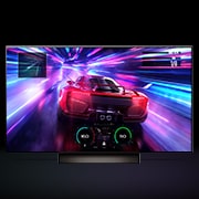 An LG C4 48 inch TV in dark space on a one-pole stand shows a colorful scene of a car racing game, and the colors reflect on the space below.