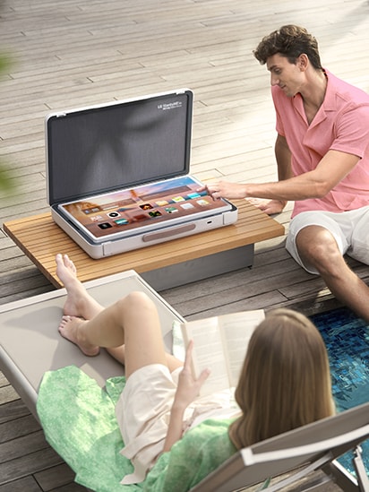 StandbyME Go se coloca sobre la mesa del patio y la pantalla se configura en modo mesa la pantalla de inicio. Un hombre está a punto de tocar una de las aplicaciones, mientras una mujer se relaja.