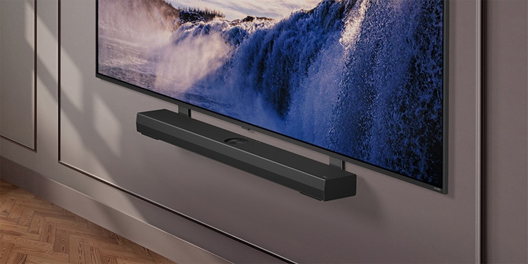 LG TV aparece con un soporte de sinergia. El soporte de sinergia y el televisor LG están conectados. La cámara hace un acercamiento al soporte, revelando la barra de sonido, que está colocada encima de él, seguida por el fondo de un espacio habitable moderno.