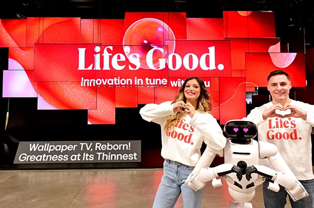 A man, a woman, and a small robot standing together in front of a red "Life's Good" display, all making heart shapes with their hands.