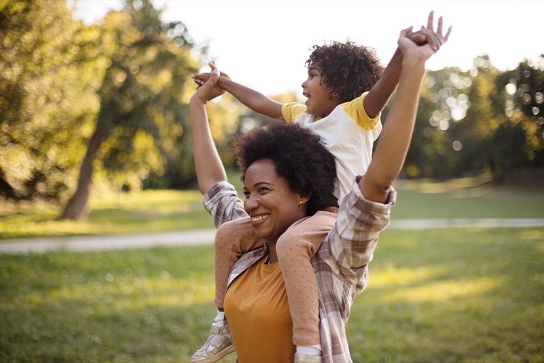 Madre e hija afroamericanas de pie en un parque. Niña sobre los hombros de la madre