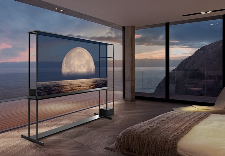 El LG SIGNATURE OLED T muestra una luna sobre la costa en su pantalla transparente. El LG SIGNATURE OLED T se encuentra en el centro de una habitación con vistas a un acantilado que da al mar. A la derecha hay una cama y un sillón.