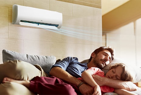 A father and daughter sleep on a couch beneath an air conditioner that is blowing air out over them.