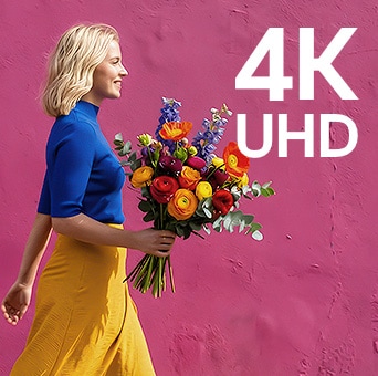 LG StanbyME 2 Max LX6 Movable Screen mit 4K UHD zeigt eine Frau in bunten Kleidung, die Blumen hält und an einer lila Straßenmauer vorbeigeht, was eine scharfe und detaillierte Auflösung hervorhebt.