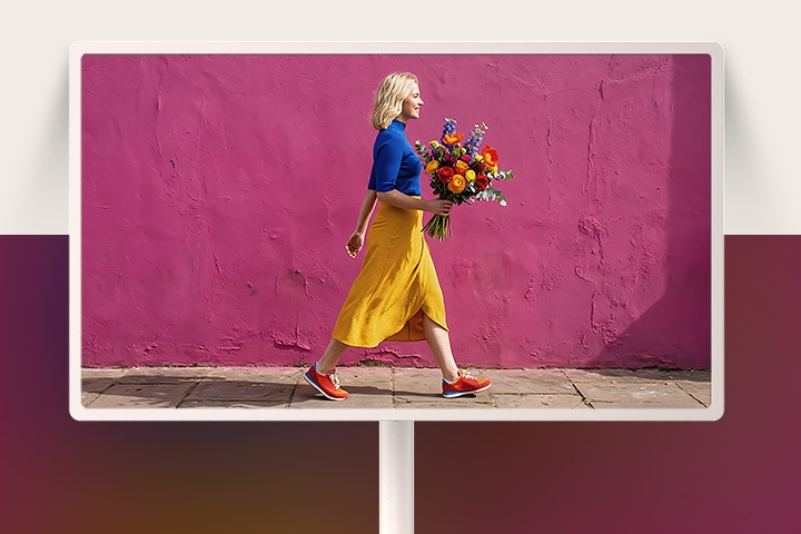 LG StanbyME 2 Max LX6 Movable Screen mit 4K UHD zeigt eine Frau in bunten Kleidung, die Blumen hält und an einer lila Straßenmauer vorbeigeht, was KI-optimierte Klarheit und verfeinerte visuelle Details hervorhebt.