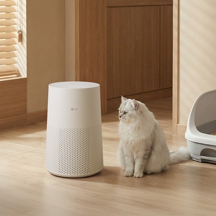 A white LG air purifier stands on a wooden floor next to a fluffy white cat and a grey litter box in a sunlit room with wooden accents