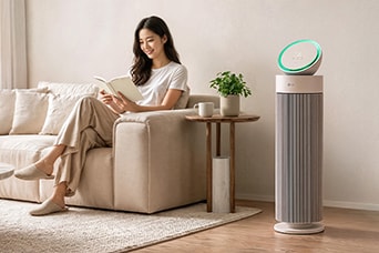 A woman reading a book on a sofa next to a tower air purifier with a glowing green light in a cozy living room.