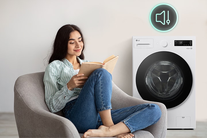 There is a washing machine in the living room and a woman is sitting in a chair reading a book