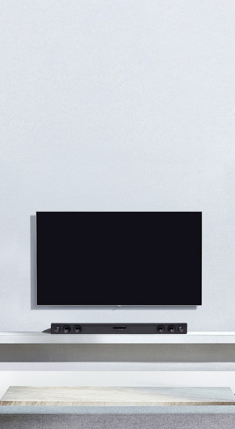LG TV is hung on the grey wall. And LG Sound Bar SQC2 is placed on the shelf in the living room. And the subwoofer is placed next to the shelf. The image illustrates that the sound bar matches the LG TV and complements the interior.