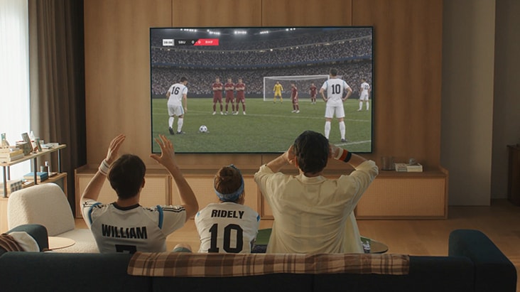LG AI TV with Sports Portal shows three people seated in a living room watching a live soccer match on a large wall-mounted TV screen during a free kick scene in a stadium. LG AI TV with Sports Portal shows three people seated in a living room watching a live soccer match on a large wall-mounted TV screen during a free kick scene in a stadium.