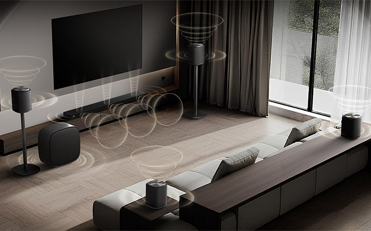LG TV with Sound Suite is wall-mounted in a modern living room, with multiple speakers arranged around a sofa as visualized sound waves radiate outward, illustrating a flexible audio setup tailored to the space.