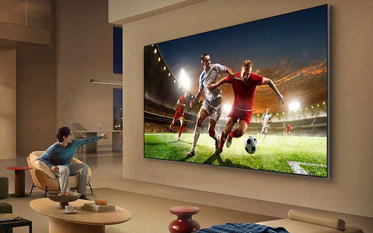 LG TV for sports viewing is wall-mounted in a spacious living room, showing a soccer match with players in motion, while a seated person points at the screen, illustrating enhanced depth and a more immersive sense of action.