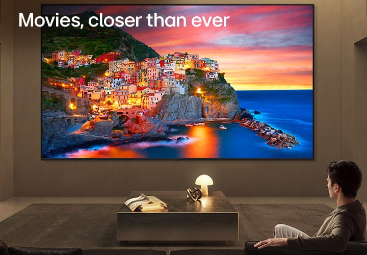 LG Ultra Big TV is wall-mounted in a spacious living room, where an expansive display shows a vibrant coastal village at sunset, illustrating how a larger screen delivers an immersive and cinematic viewing experience.